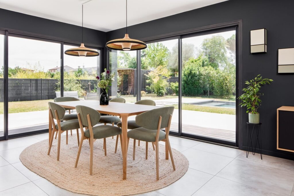 Salle à manger avec chaises vertes et tapis en jute, baies vitrées ouvertes sur le jardin et la piscine