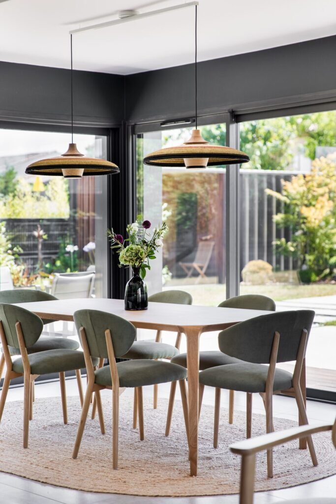 Table à manger en bois, chaises vertes et tapis en jute rond devant les baies vitrées